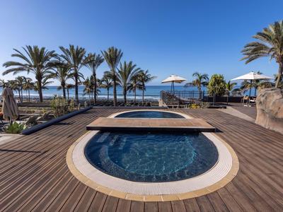 Piscine avec terrasse en bois, palmiers et vue sur la mer sous un ciel dégagé.