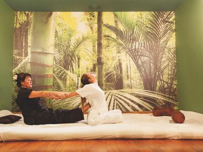 Two people practicing a partner stretch on a mat in a room with green walls and tropical mural.