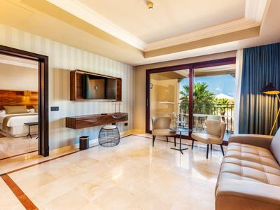 Bright hotel room with seating area, TV, and balcony overlooking palm trees.