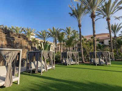 Palm trees and cabanas on a green lawn beside rocky hills under a blue sky.