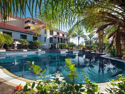 Hotel met groot buitenzwembad, omgeven door palmbomen en groen bij zonnig weer.