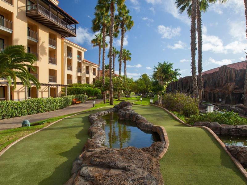 Resort building with tall palm trees and a small water stream on green grass under blue sky.