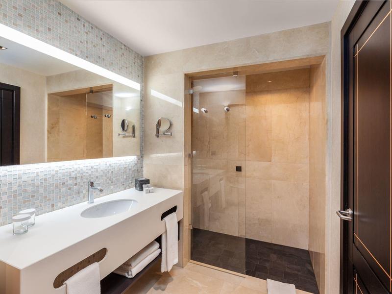 Modern hotel bathroom with double sink, mirror, towels, and walk-in shower with glass door.