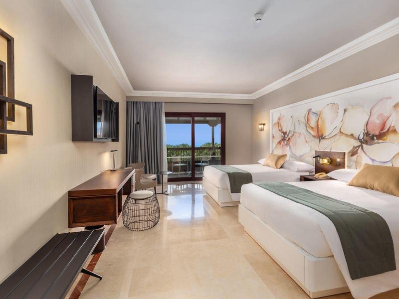 Bright hotel room with two beds, large art above headboards, balcony view, and modern furnishings.