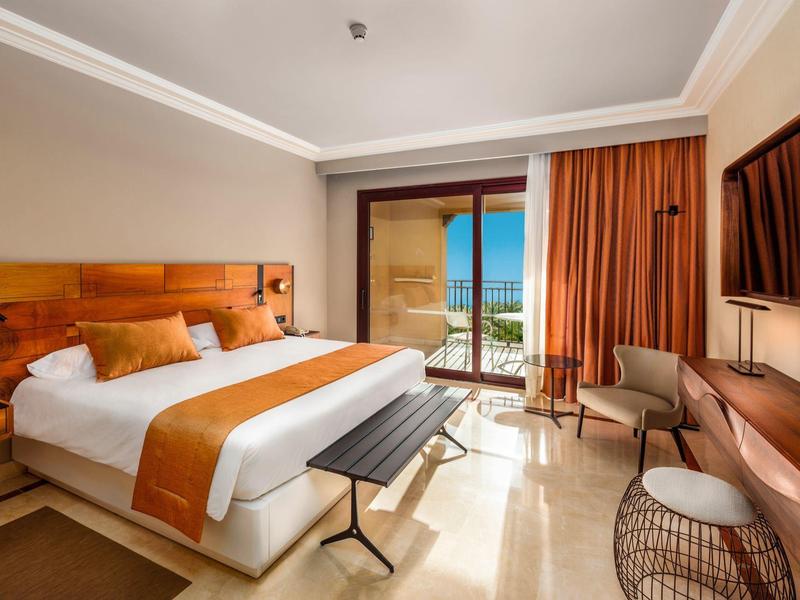 Bright hotel room with king bed, wooden headboard, balcony, sea view, and modern furnishings.