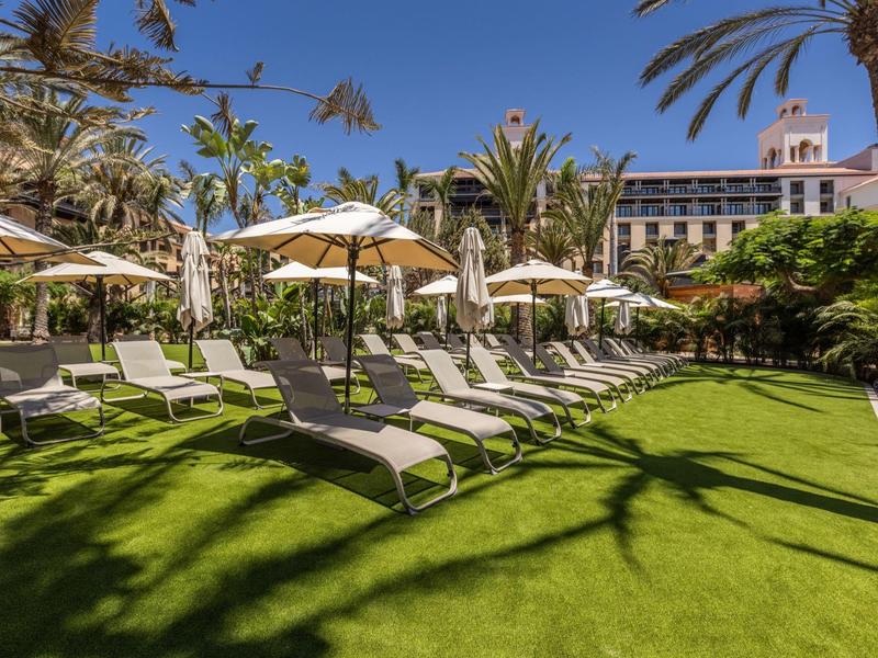 Rows of lounge chairs under umbrellas on green grass near palm trees and a hotel building.
