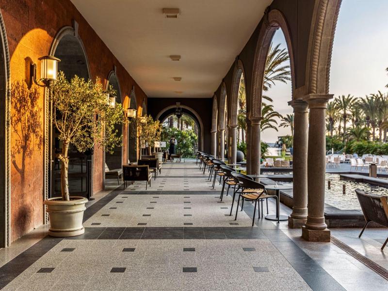 Arched outdoor corridor with seating and potted plants by a pool under evening light.