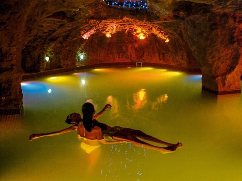 Person floating on inflatable in dimly lit cave pool with warm yellow lights.