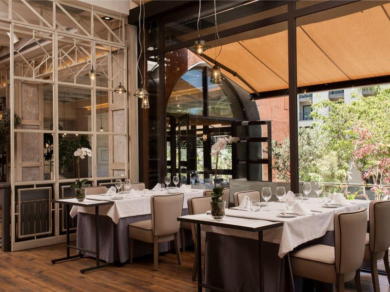 Restaurante elegante con mesas preparadas, grandes ventanales y vista a un área exterior con vegetación.