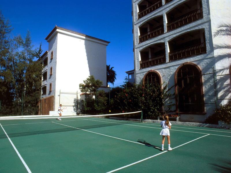 Děti hrají tenis na kurtu vedle hotelové budovy za slunečného počasí.