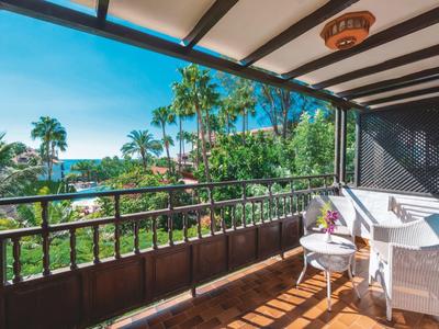 Balkon met stoelen en tafel met uitzicht op palmbomen en zee op een zonnige dag.
