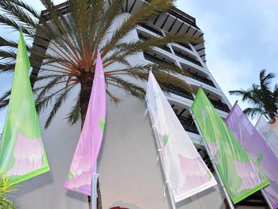 Kleurrijke vlaggen wapperen voor een hoog gebouw met palmbomen onder een bewolkte hemel.