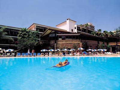 View of a pool with a person on an air mattress in front of a multi-story hotel building and sun umbrellas.