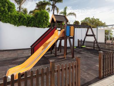 Parco giochi per bambini con scivolo e altalena su pavimento lastricato, circondato da recinzione in legno.