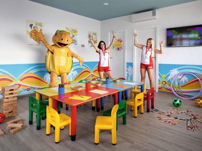 Colorful children's playroom with tables, chairs, toys, and three people, one in a yellow costume.