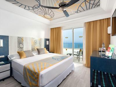Modern hotel room with double bed, ceiling fan, and balcony overlooking the sea.