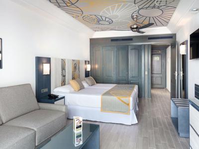 Bright hotel room with two beds, sofa, ceiling fan, and modern decor.