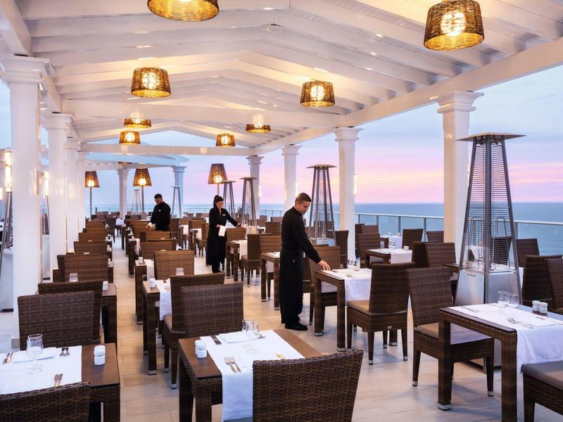 Elegant restaurant with set tables, wicker chairs, and a sea sunset view.