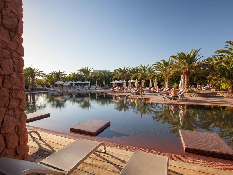 Piscina all'aperto circondata da palme con lettini e muri di pietra rossa.