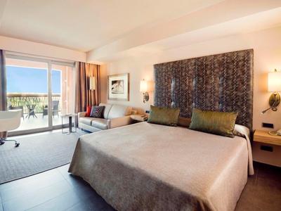 Bright hotel room with large bed, sofa, and balcony overlooking greenery and water.