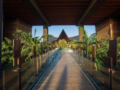 Wooden walkway with glass railings leading to tropical garden under a wooden roof.