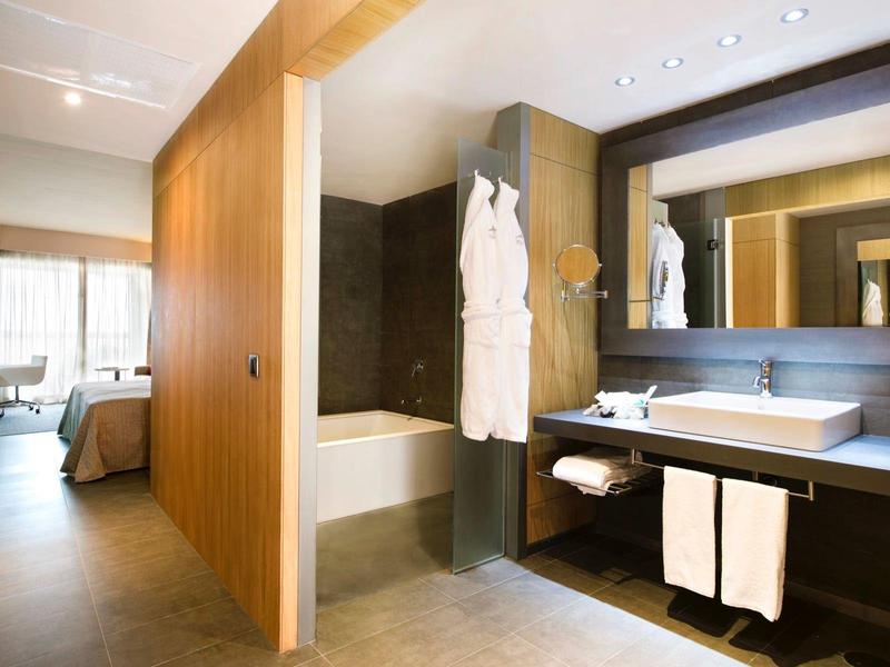 Modern hotel bathroom with bathtub, dark tiles, wooden panels, and a connected bedroom.