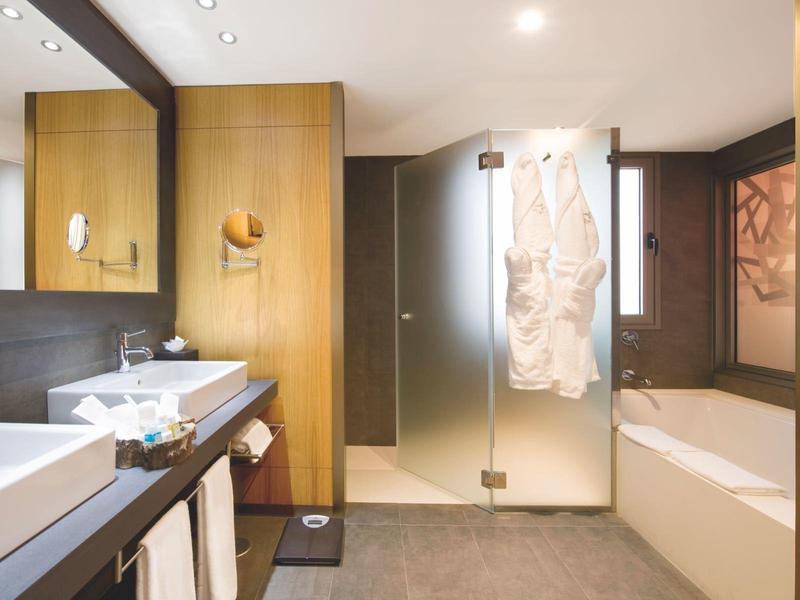 Modern hotel bathroom with double sinks, glass shower, bathtub, wooden panels, and bathrobes.