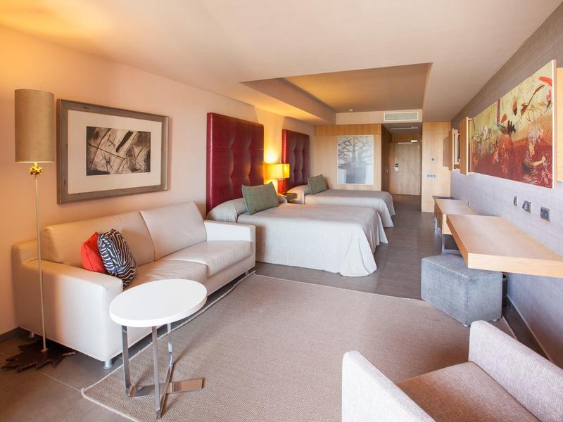 Spacious hotel room with two beds, sofa, armchair, desk, and warm lighting.