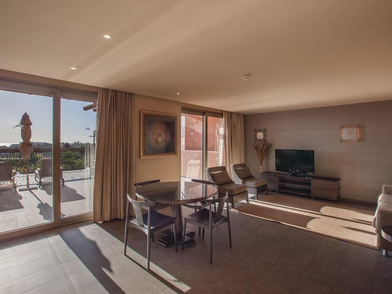 Bright hotel suite with dining table, seating area, and balcony with outdoor view.