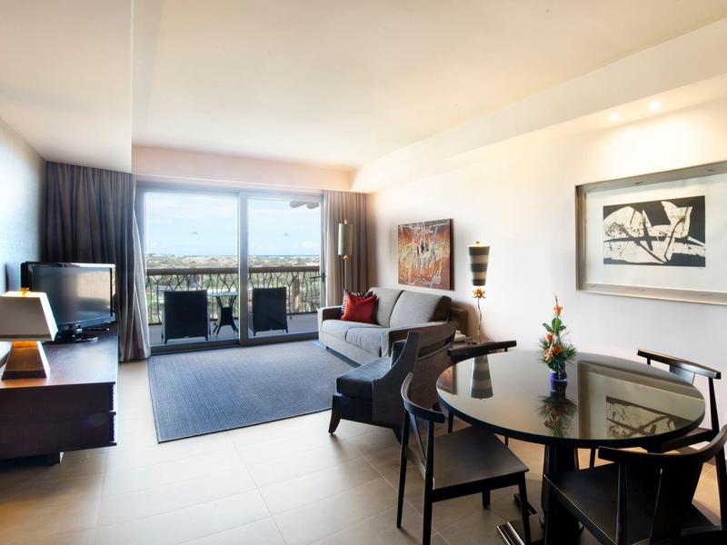 Bright hotel room with glass table, armchair, dresser, and balcony overlooking cityscape.