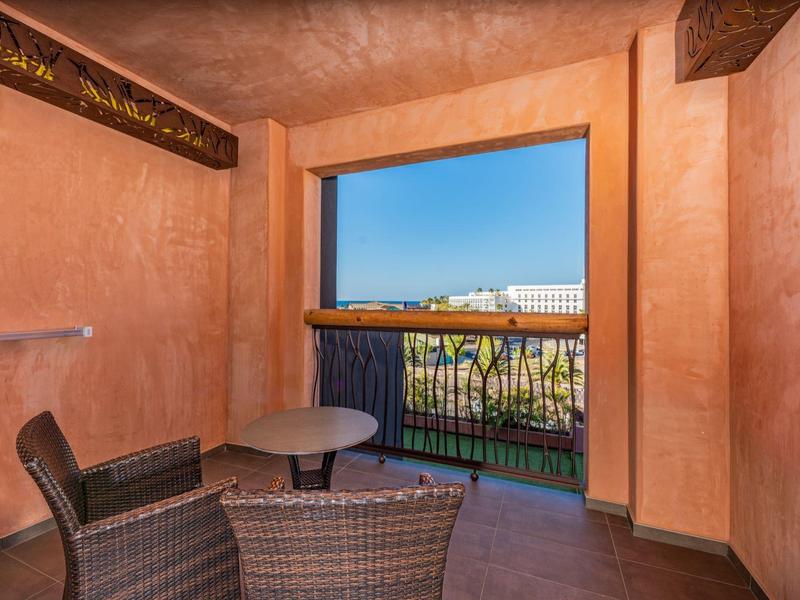Cozy balcony with wicker chairs and a table overlooking a distant hotel and palm trees under blue sky.