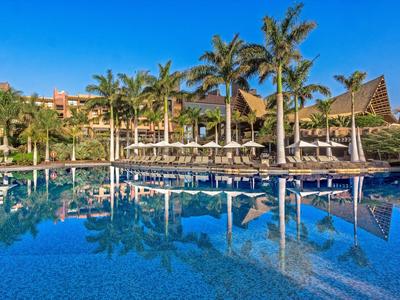 Grande piscine avec chaises longues et palmiers devant un hôtel sous un ciel bleu clair.