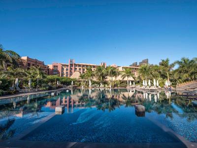 Grande piscine avec des palmiers et des bâtiments d'hôtel sous un ciel dégagé.