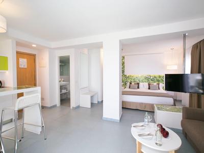 Modern hotel suite with kitchenette, dining area, seating, and bedroom separated by partial wall.