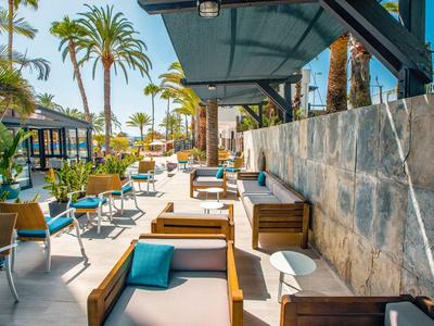 Sunny outdoor hotel patio with wooden lounge chairs, blue cushions, and palm trees.