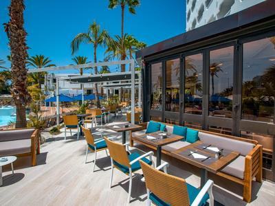Terrazza esterna dell'hotel con tavoli e sedie vicino alla piscina sotto palme in una giornata di sole.
