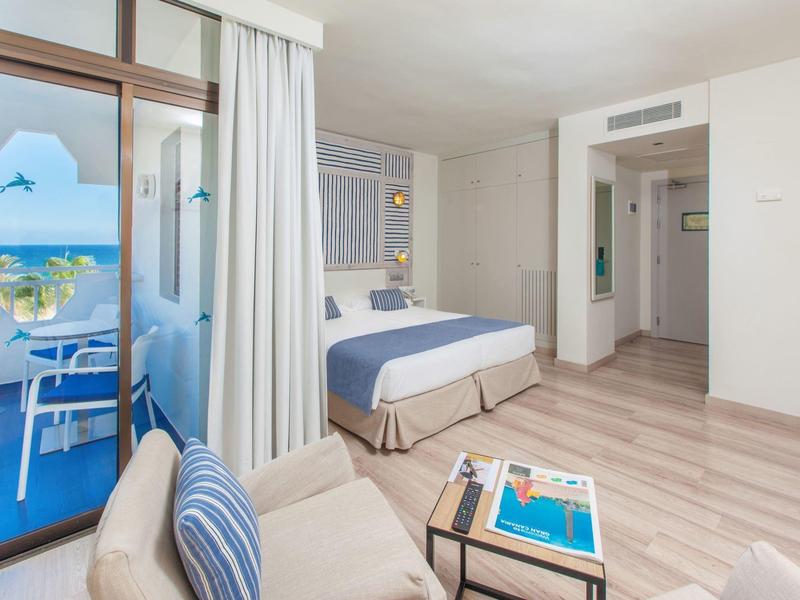 Camera d'hotel luminosa con accenti blu, balcone con vista mare, zona soggiorno accogliente e arredamento moderno.