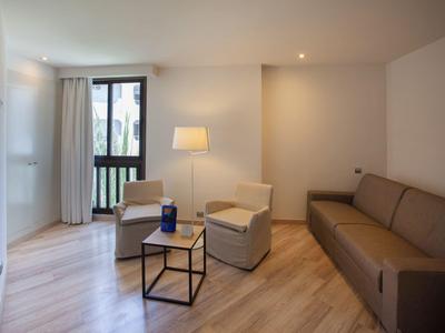 Chambre d'hôtel moderne avec fauteuils beige, canapé, table basse et grande fenêtre avec rideaux.