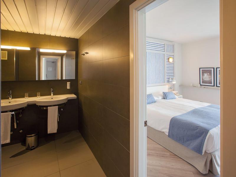 Vue d'une salle de bain avec doubles lavabos menant à une chambre lumineuse avec un lit double.