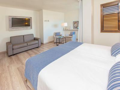 Hell eingerichtetes Schlafzimmer mit Doppelbett, Sofa, Holzboden und einem Fenster mit Jalousien.