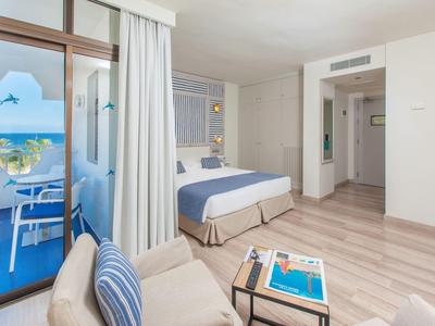 Helles Hotelzimmer mit Doppelbett, Sofa, blau-weißen Akzenten und Blick auf Balkon mit Meer.