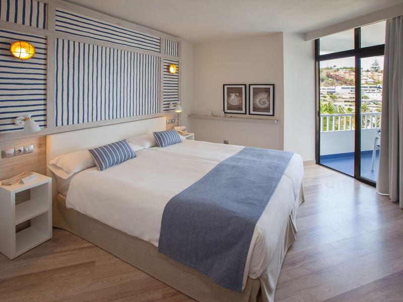 Hell eingerichtetes Schlafzimmer mit Doppelbett, blauen Kissen und Terrasse mit Meerblick.