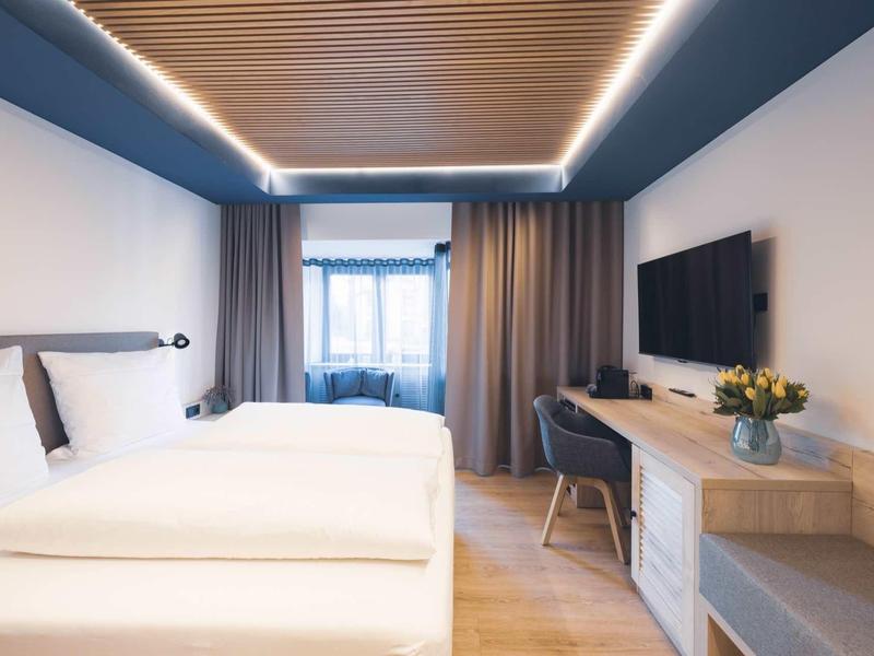 Modernes Hotelzimmer mit zwei Betten, Holzboden, großer Fensterfront und Fernseher an der Wand.