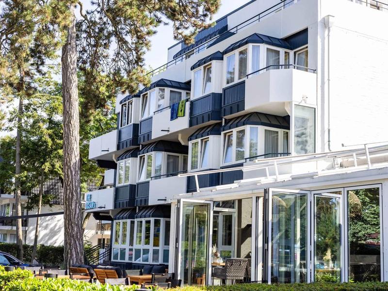 Mehrstöckiges weißes Gebäude mit Balkonen, umgeben von Bäumen und Grünflächen bei Tageslicht.
