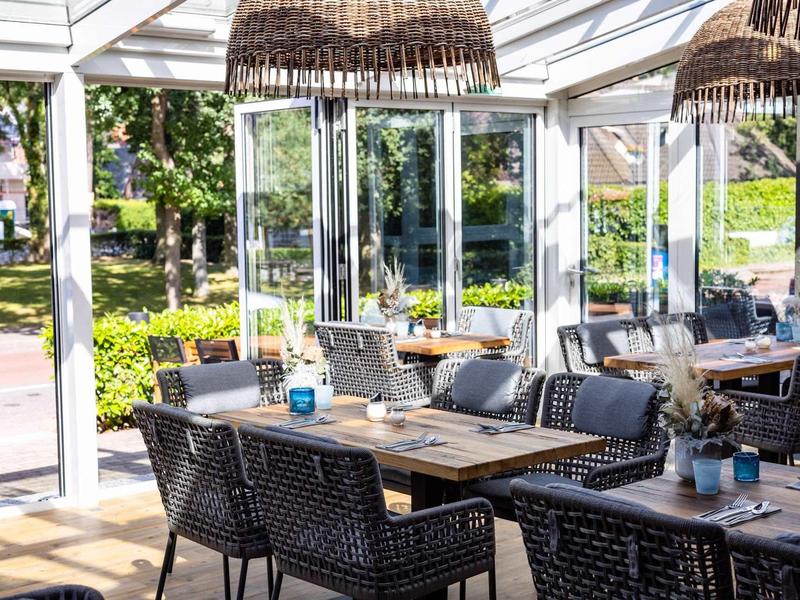 Außenterrasse mit Holztischen, Rattanstühlen und Glasdach, umgeben von grünem Garten.