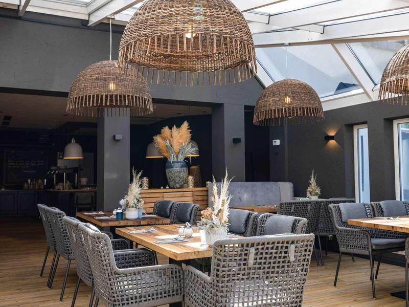 Modernes Restaurant mit runden Holztischen, grauen Stühlen und großen geflochtenen Lampenschirmen.