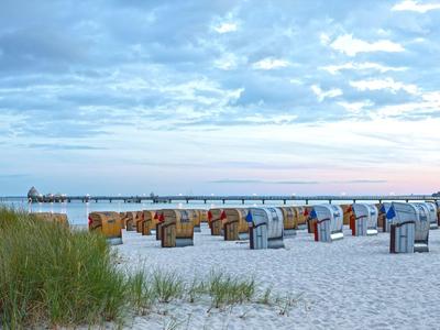 Weitläufiger Sandstrand mit Strandkörben und einem Pier im Hintergrund bei bewölktem Himmel.