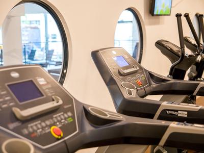 Fitnessraum mit Laufbändern und elliptischen Trainern vor runden Fenstern.