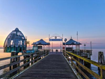 Holzsteg mit Pavillons und einer Skulptur, die ins ruhige Meer bei Sonnenuntergang führt.