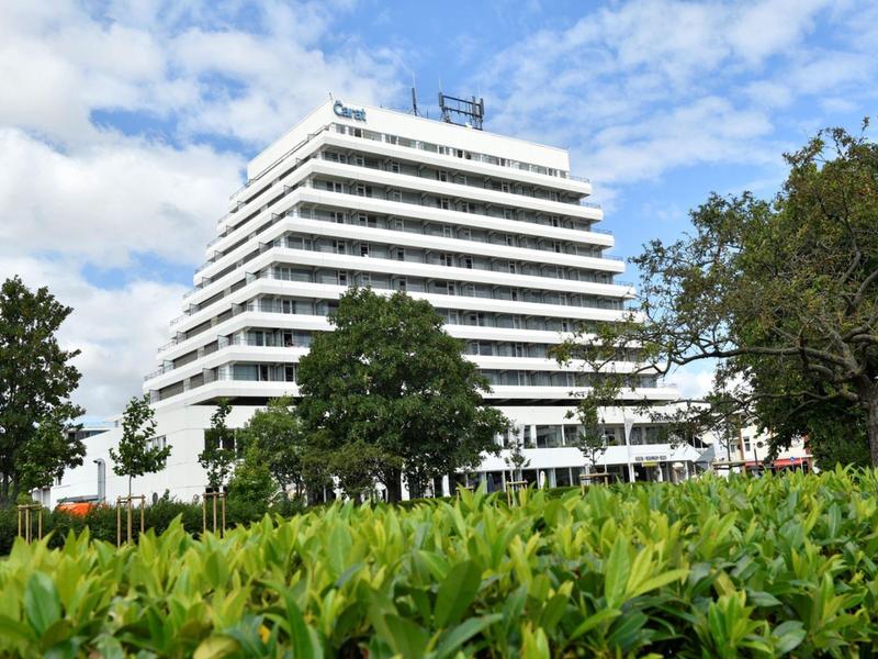 Modernes mehrstöckiges Hotelgebäude mit Balkonen, umgeben von Bäumen und einem blauen Himmel.